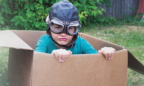 A child wearing an aviator helmet and goggles plays in a cardboard box.